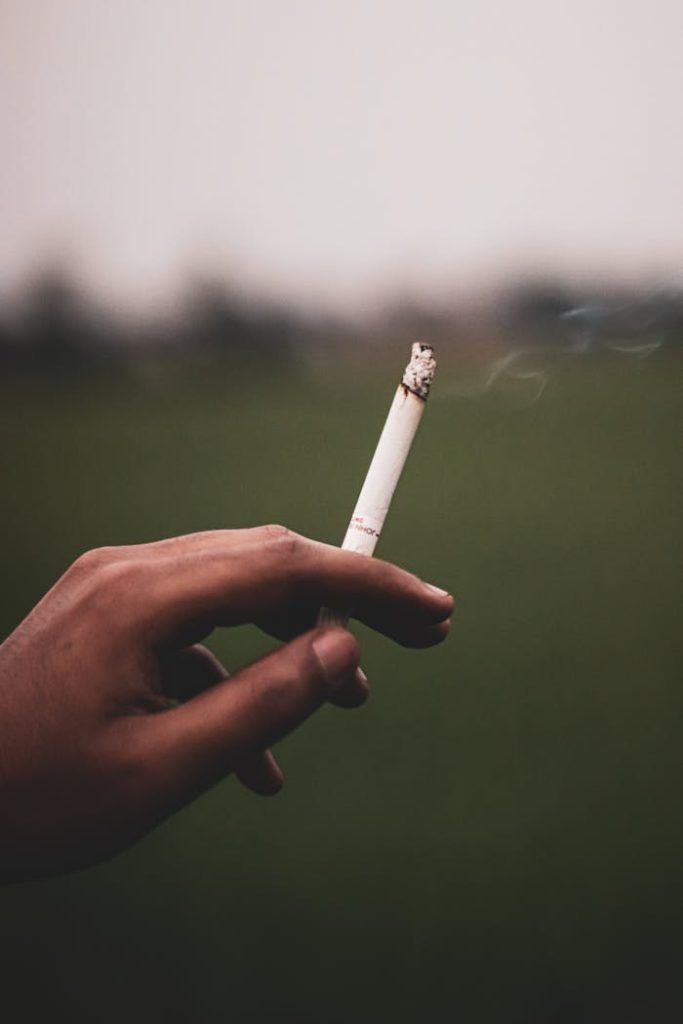 Close-up of a hand holding a lit cigarette with blurred outdoor background.
